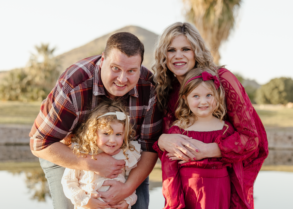 A smiling family of four poses near a pond, parents hugging their two young daughters in red dresses.