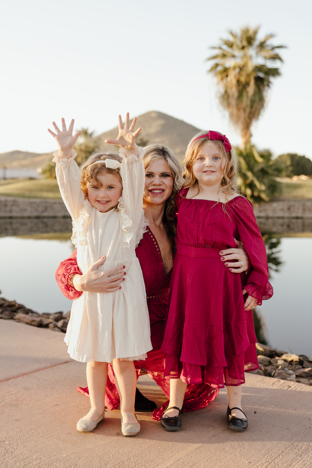 A mother kneels between her two daughters in red and white dresses near a scenic pond with palms.
