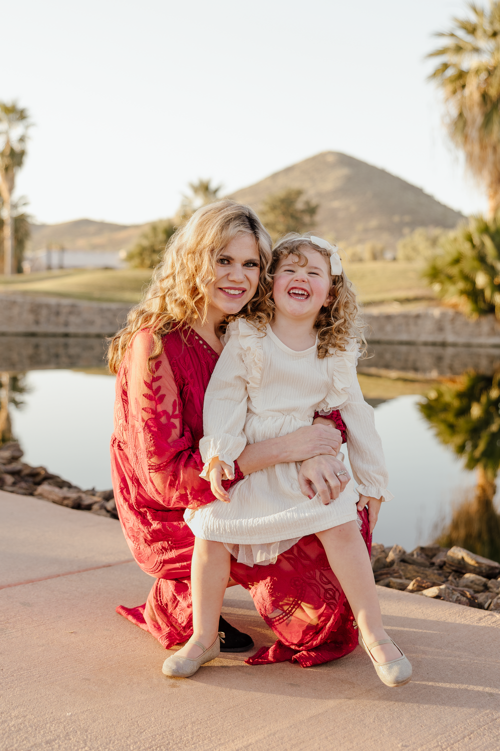 Mother in red dress kneeling with laughing curly-haired daughter in white dress by a scenic pond and mountain background.