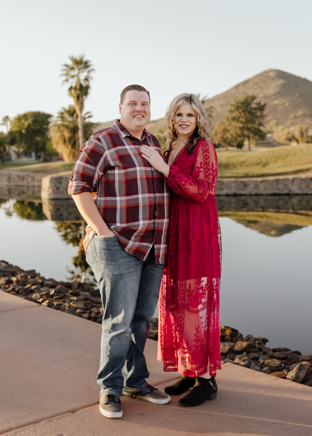 Couple posing by a reflective pond with palm trees and a mountain backdrop, woman in red lace dress, man in plaid shirt.