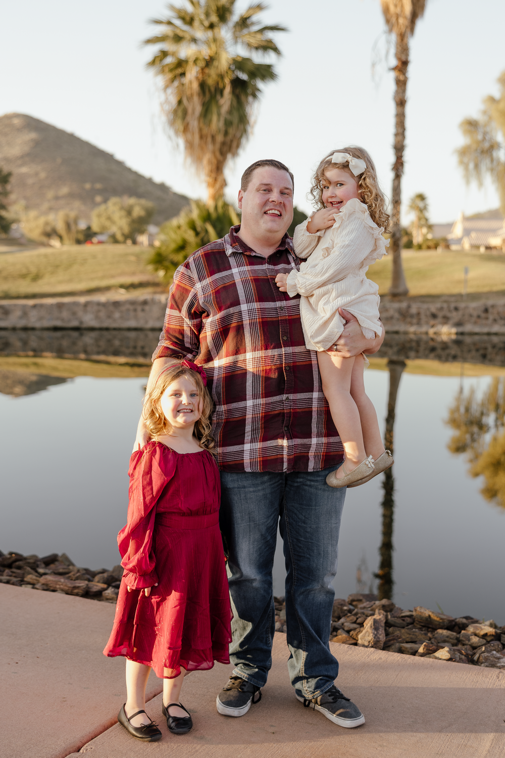 Father in plaid shirt posing with two young daughters in red and white dresses by a reflective pond outdoors.
