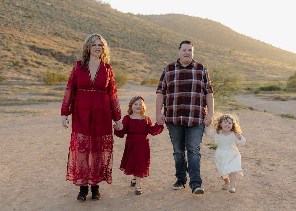 Family of four walking hand-in-hand in a desert landscape at sunset, wearing coordinated red and plaid outfits.