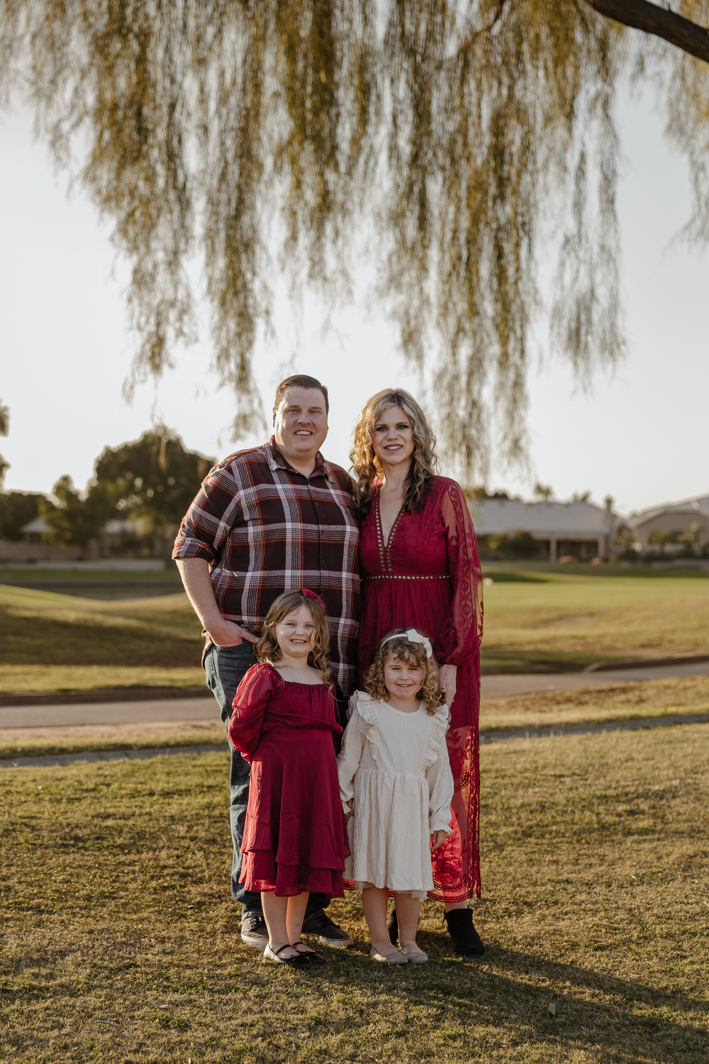 A family of four poses under a willow tree, parents and two young daughters in red and white dresses.