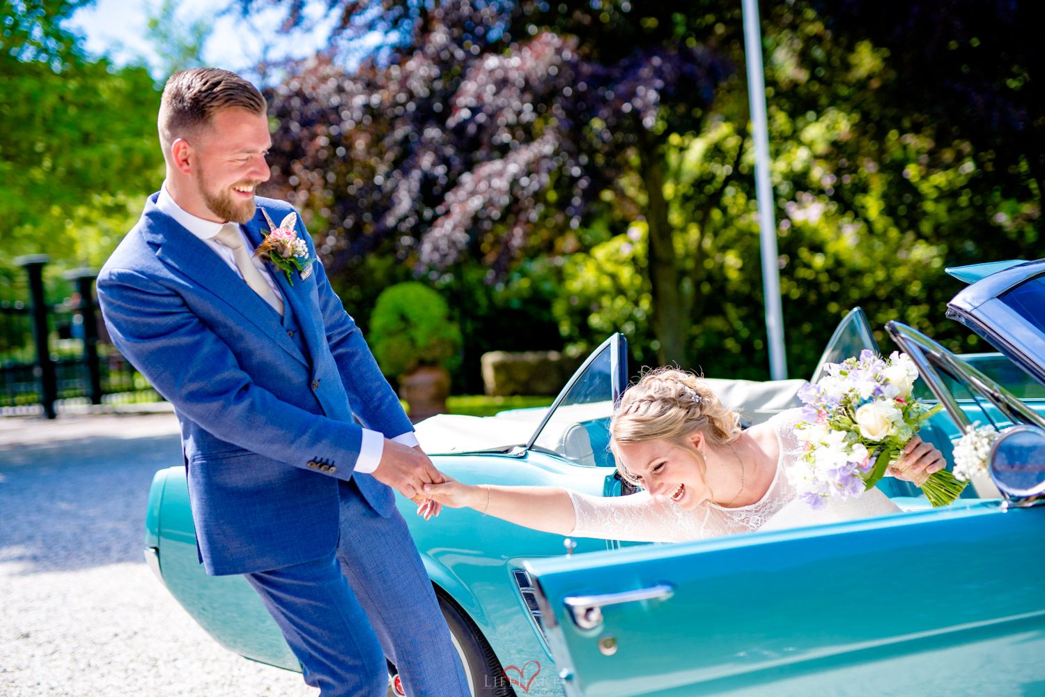Person in bright blue suit poses playfully next to a turquoise classic car decorated with flowers at an outdoor event.