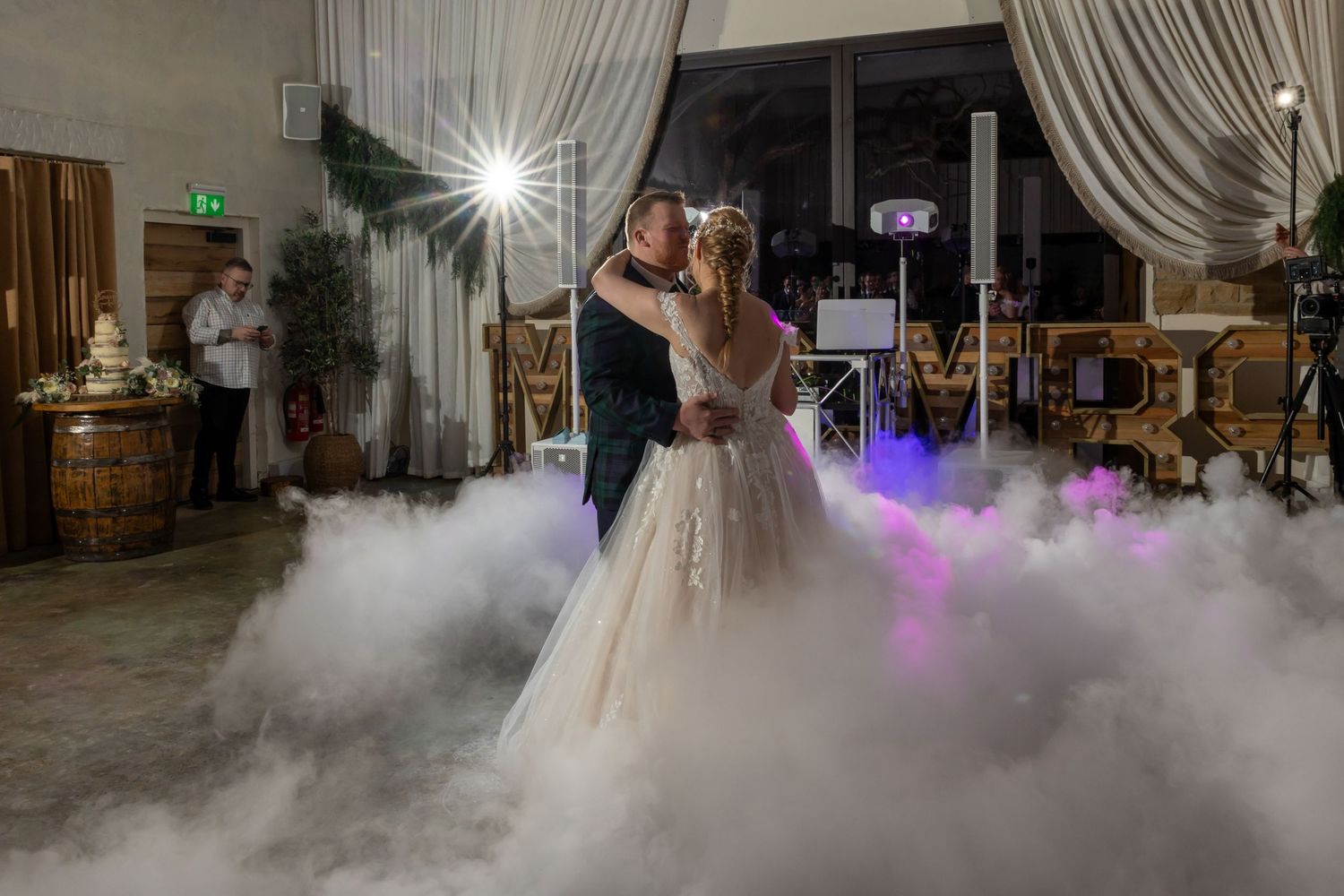 A wedding couple dances on a fog-covered dance floor with purple lighting and white curtains in the background.