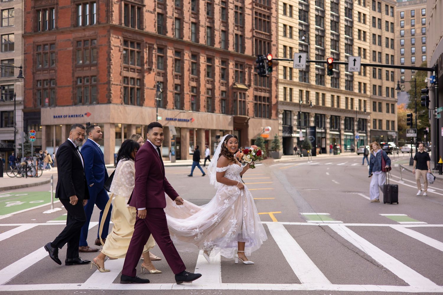 A wedding party crosses a busy intersection in New York City with tall historic buildings in the background.