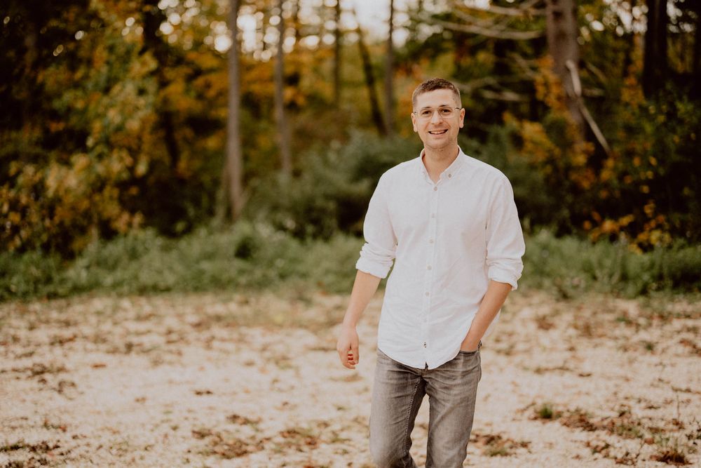 Person in white button-down shirt and gray pants standing on sandy ground with autumn forest in background.