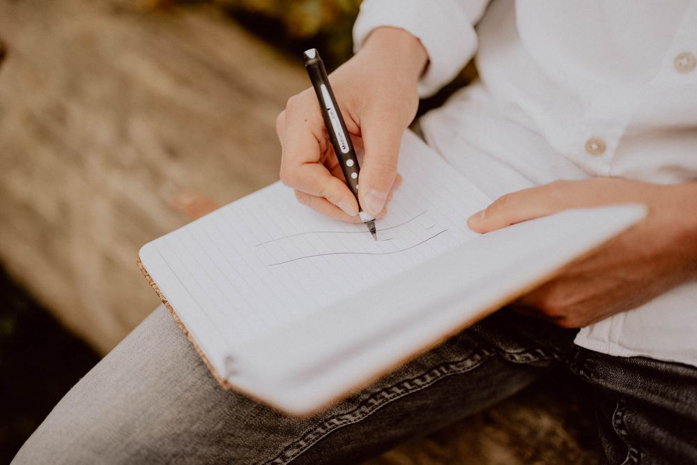 Hands holding a pen and writing in a white notebook on a rustic wooden surface.