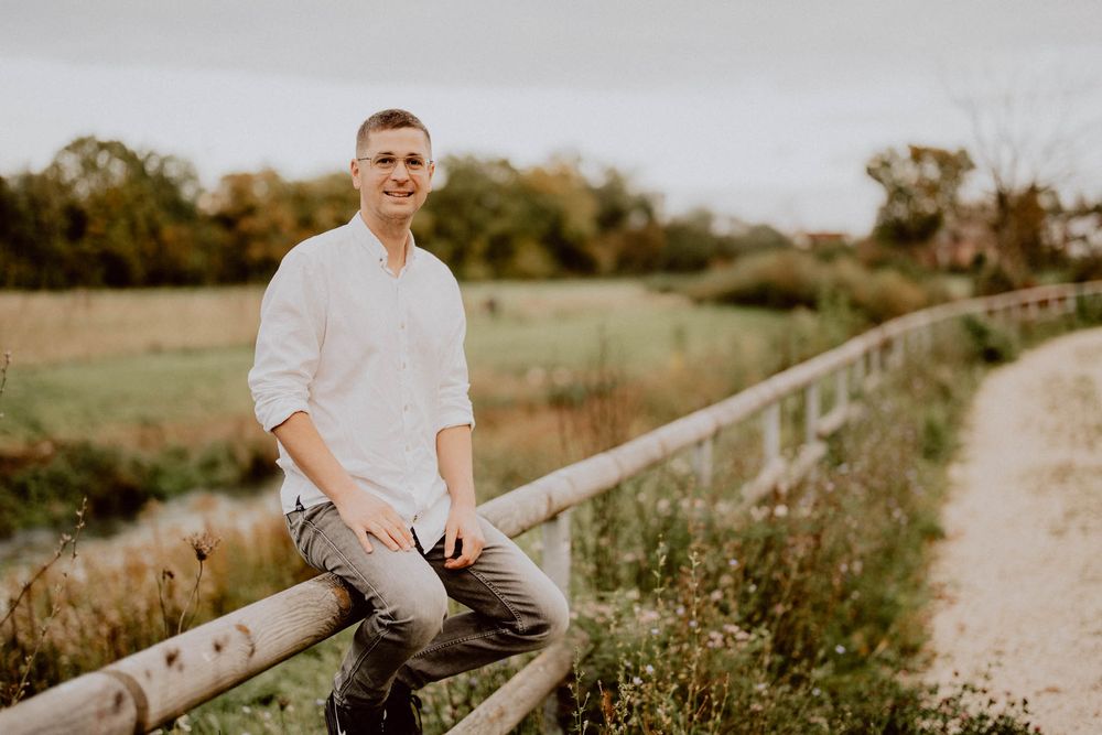 A person in a white shirt sits casually on a wooden fence along a rural dirt path surrounded by greenery.