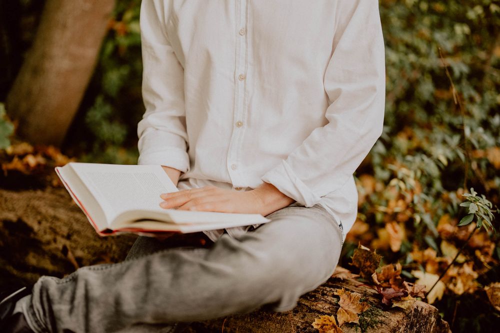 A person in white clothing sits outdoors reading a book against an autumn backdrop with fallen leaves.