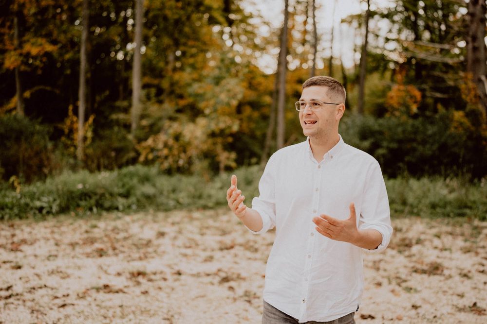 A person in a white button-down shirt gestures while standing in a sunlit clearing surrounded by autumn trees.