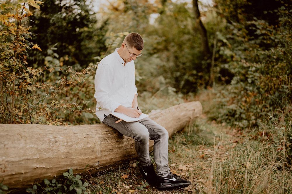 Someone sitting on a fallen log writing in a notebook outdoors.