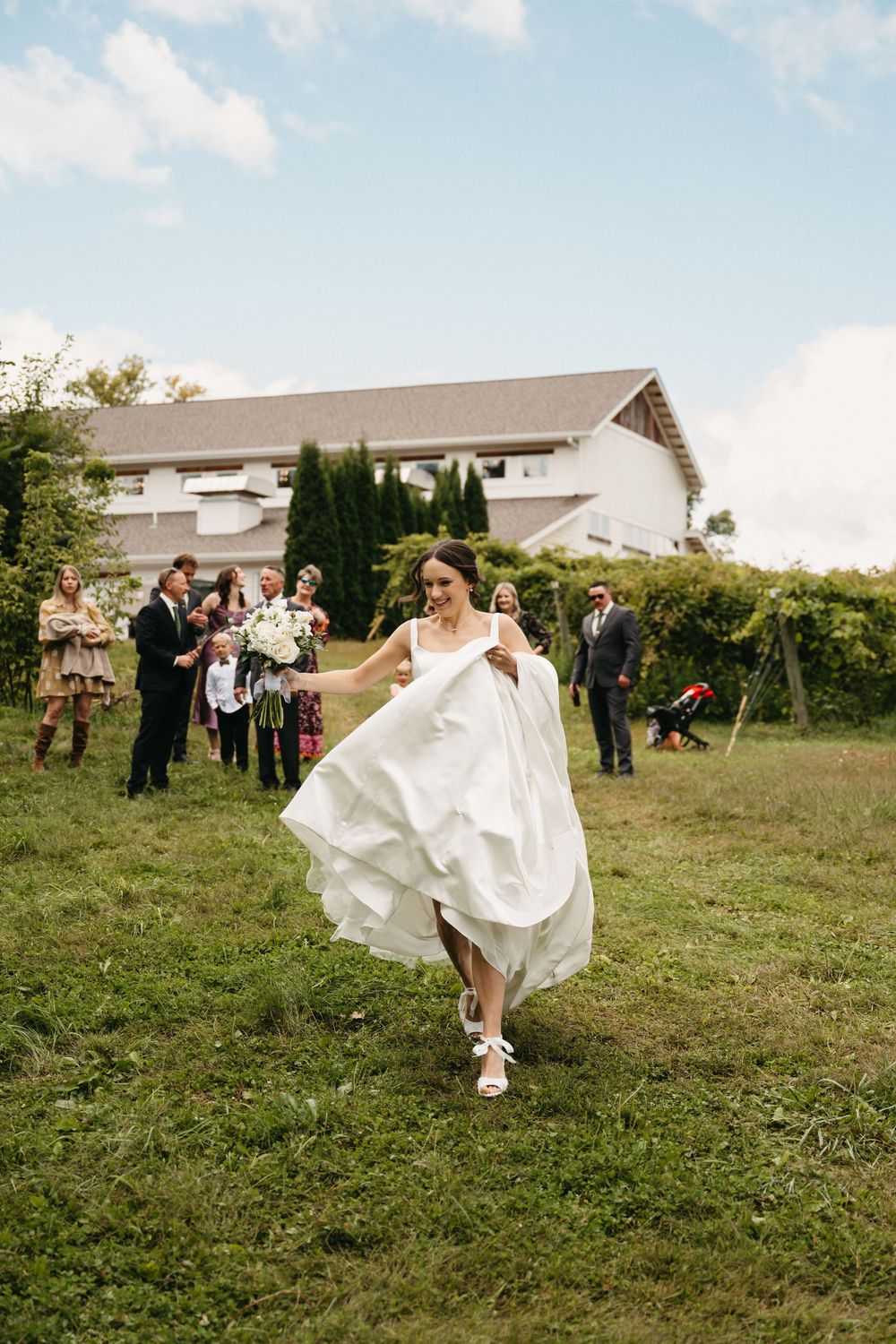 A joyful scene unfolds as a white wedding dress billows in the breeze on a grassy hillside with guests watching.