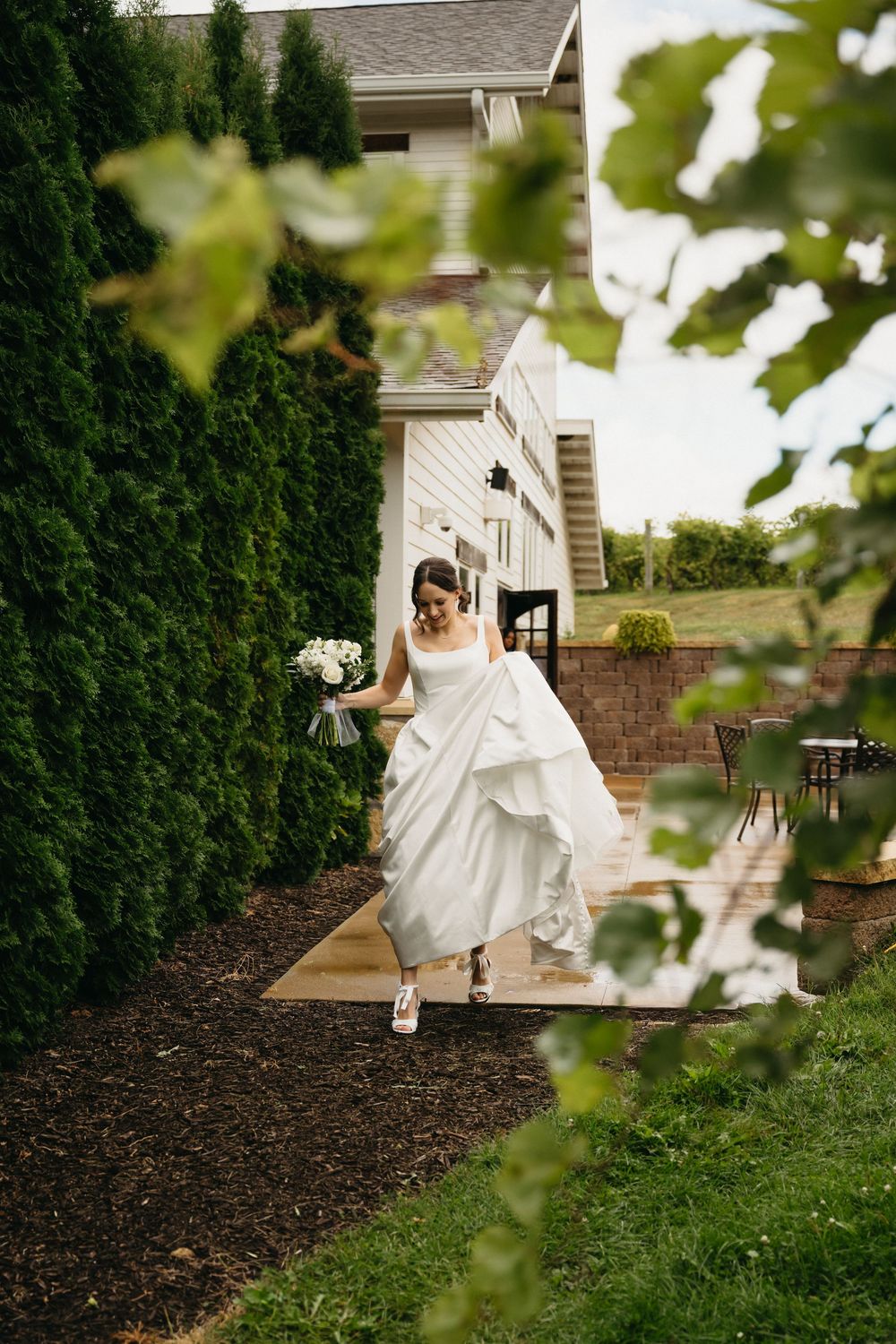 A flowing white wedding dress twirls gracefully against a backdrop of green hedges and a house exterior.
