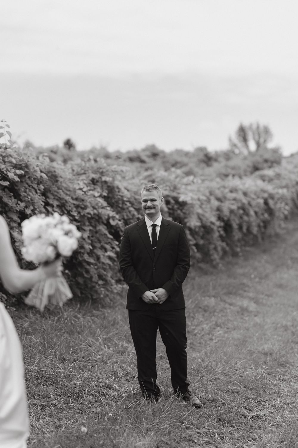 A sequence of black and white photos showing someone in a dark suit standing in a grassy field with hedges.
