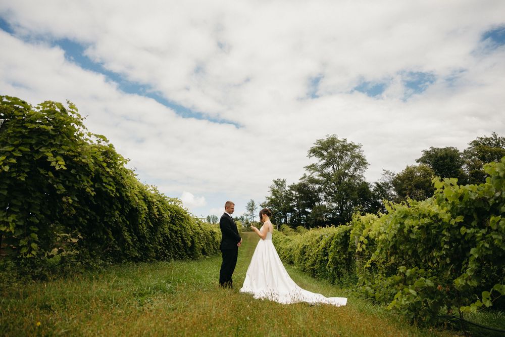 A couple stands between lush green hedgerows on their wedding day under a bright cloudy sky.