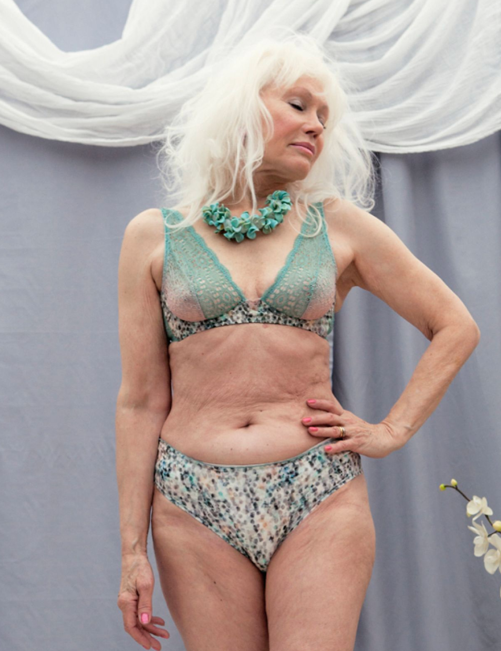 Woman in teal lingerie and statement necklace posing against draped fabric background.