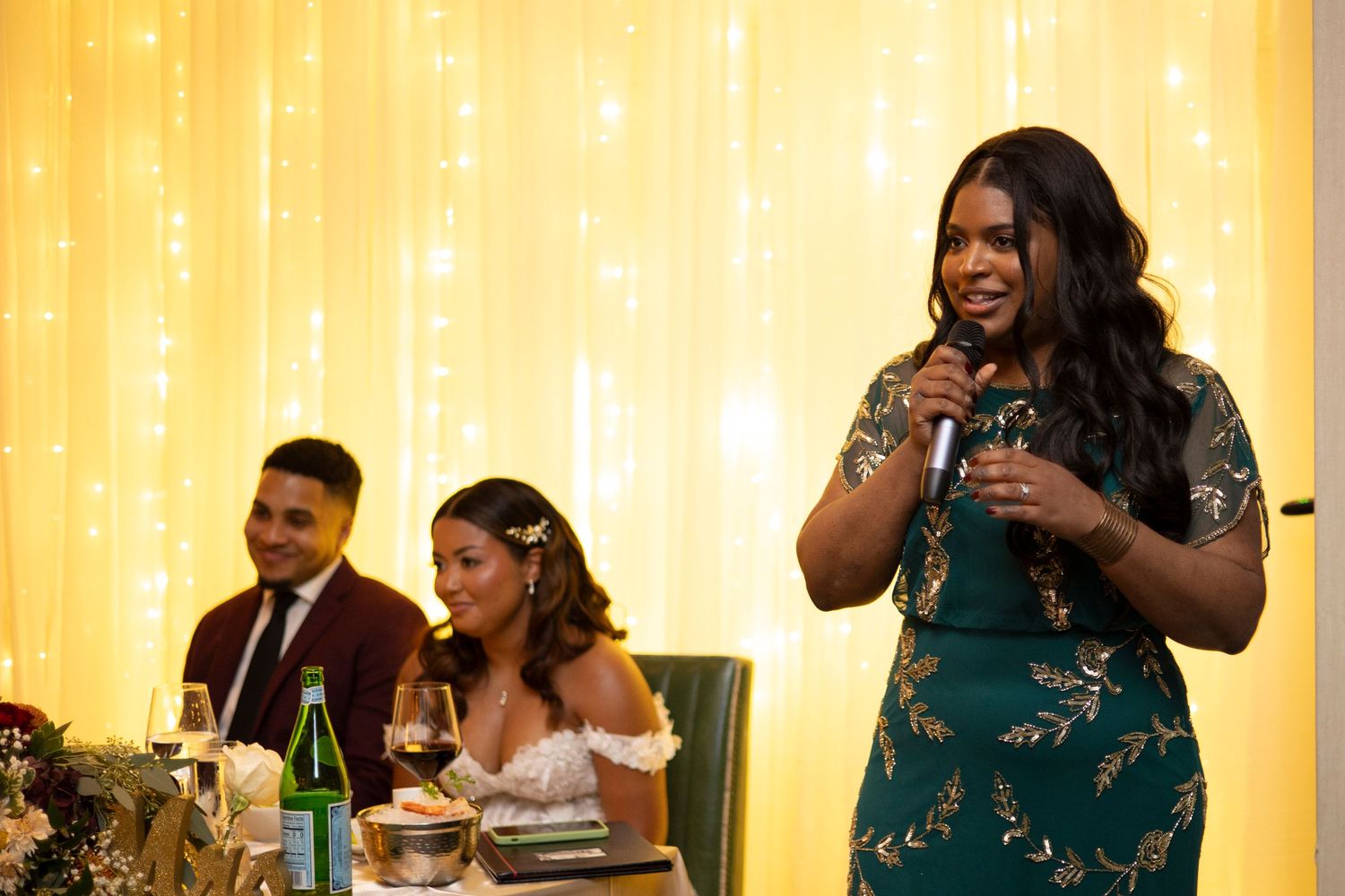 Wedding reception guests gather at tables in a warmly lit banquet hall with yellow drapes as backdrop.