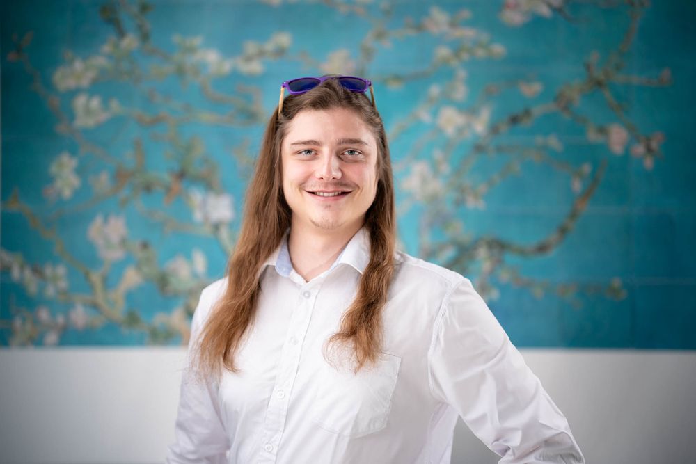Professional headshot portrait against a turquoise textured wall background wearing a white button-up shirt.