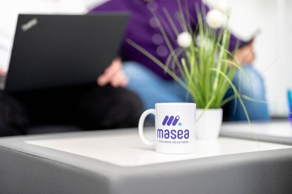 A white coffee mug with a purple logo sits on a gray surface next to a laptop and decorative plant.