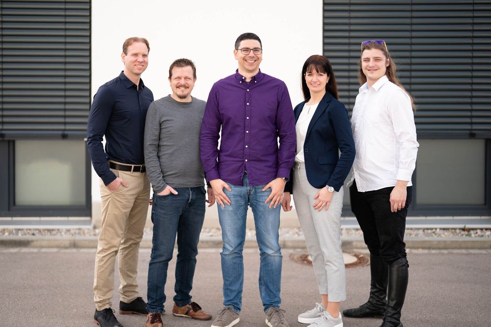 A group of five people standing in casual business attire against a modern gray building exterior.
