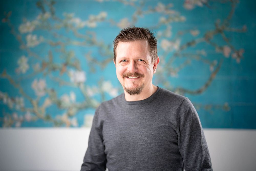 Professional headshot portrait against a turquoise and gray background showing a confident smile and gray sweater.