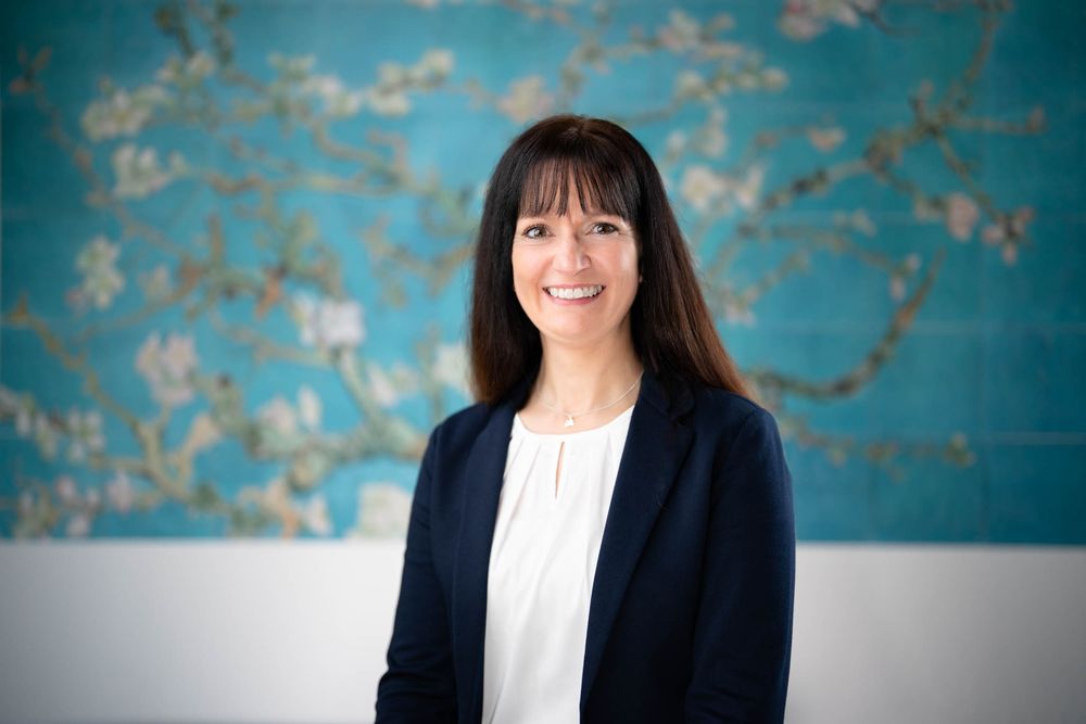 Professional business portrait against a teal textured wall with dark blazer and white blouse.