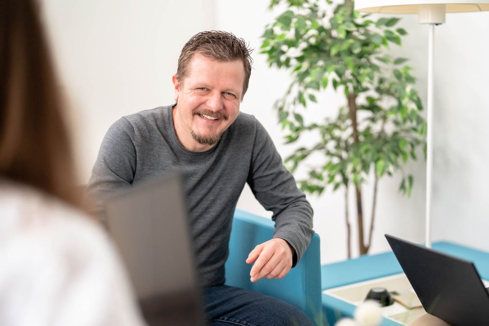 A person in a gray sweater smiles while sitting at a desk in a bright modern office setting.
