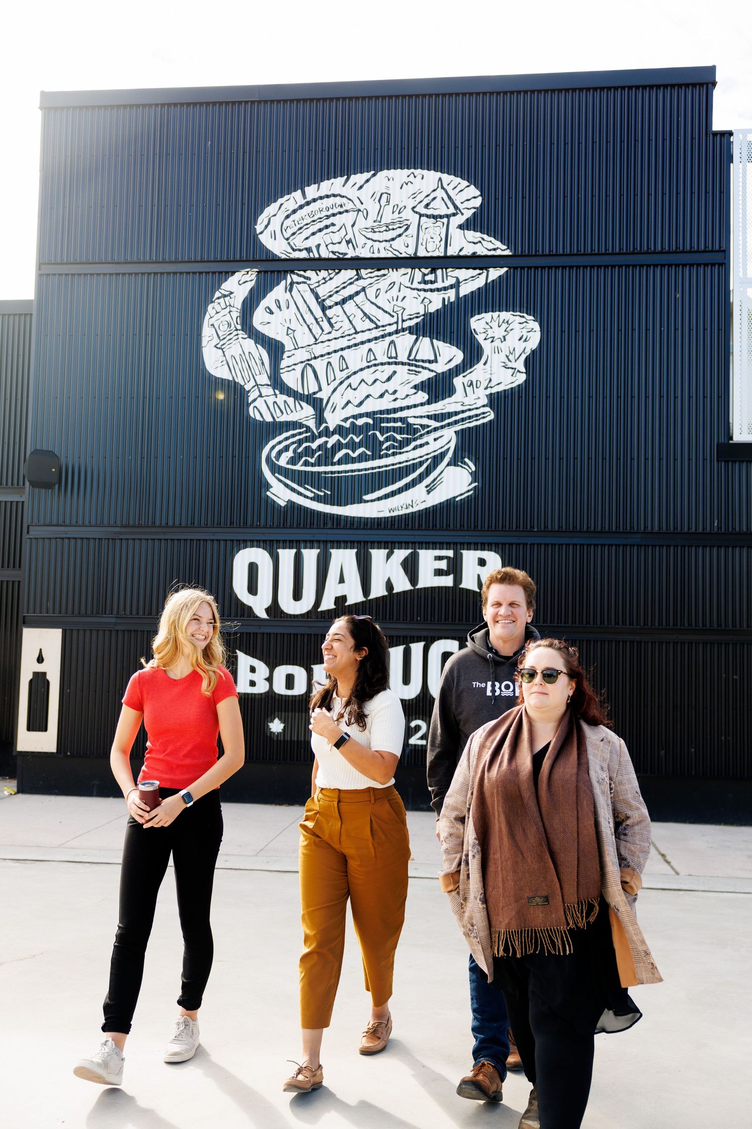 A group stands in front of a black wall with a large white graphic design and Quaker logo at a business location.