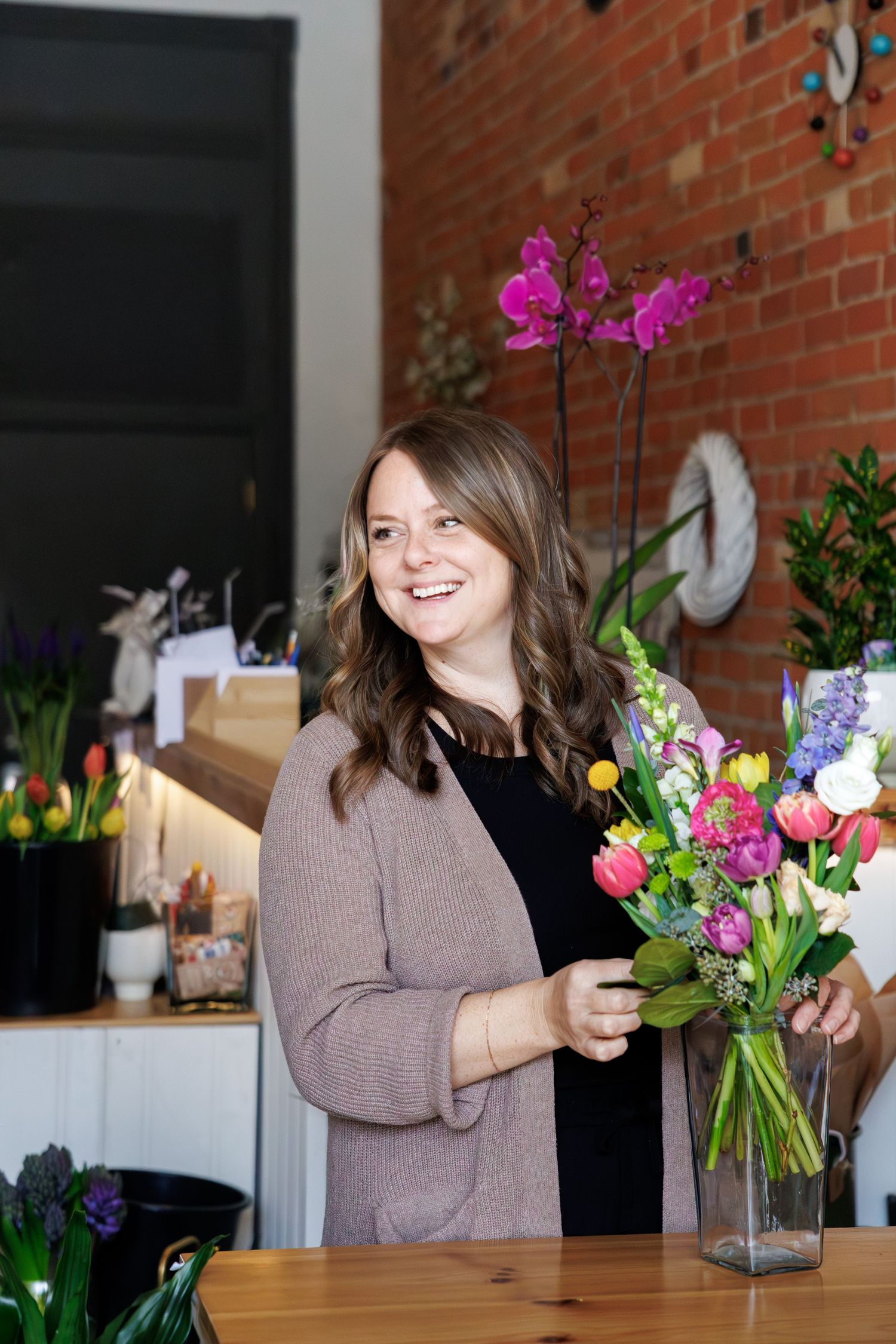 Smiling florist arranging colorful spring bouquet with tulips and orchids in a glass vase at a flower shop.