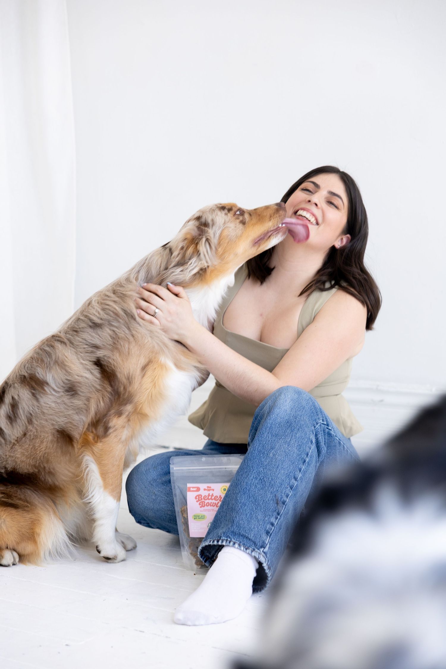 A happy woman sits on the floor laughing as her Australian Shepherd dog licks her face, with a pet treat bag nearby.