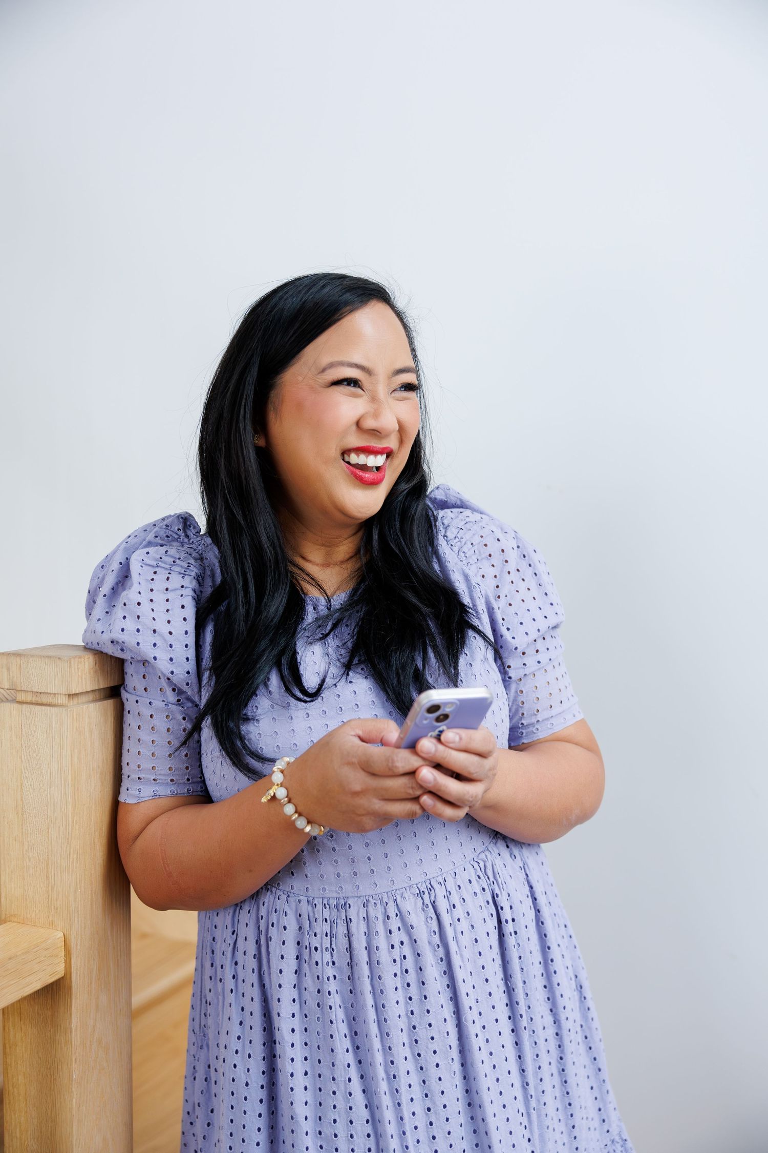 Woman in lavender crochet dress laughs joyfully while looking at mobile phone against white background.