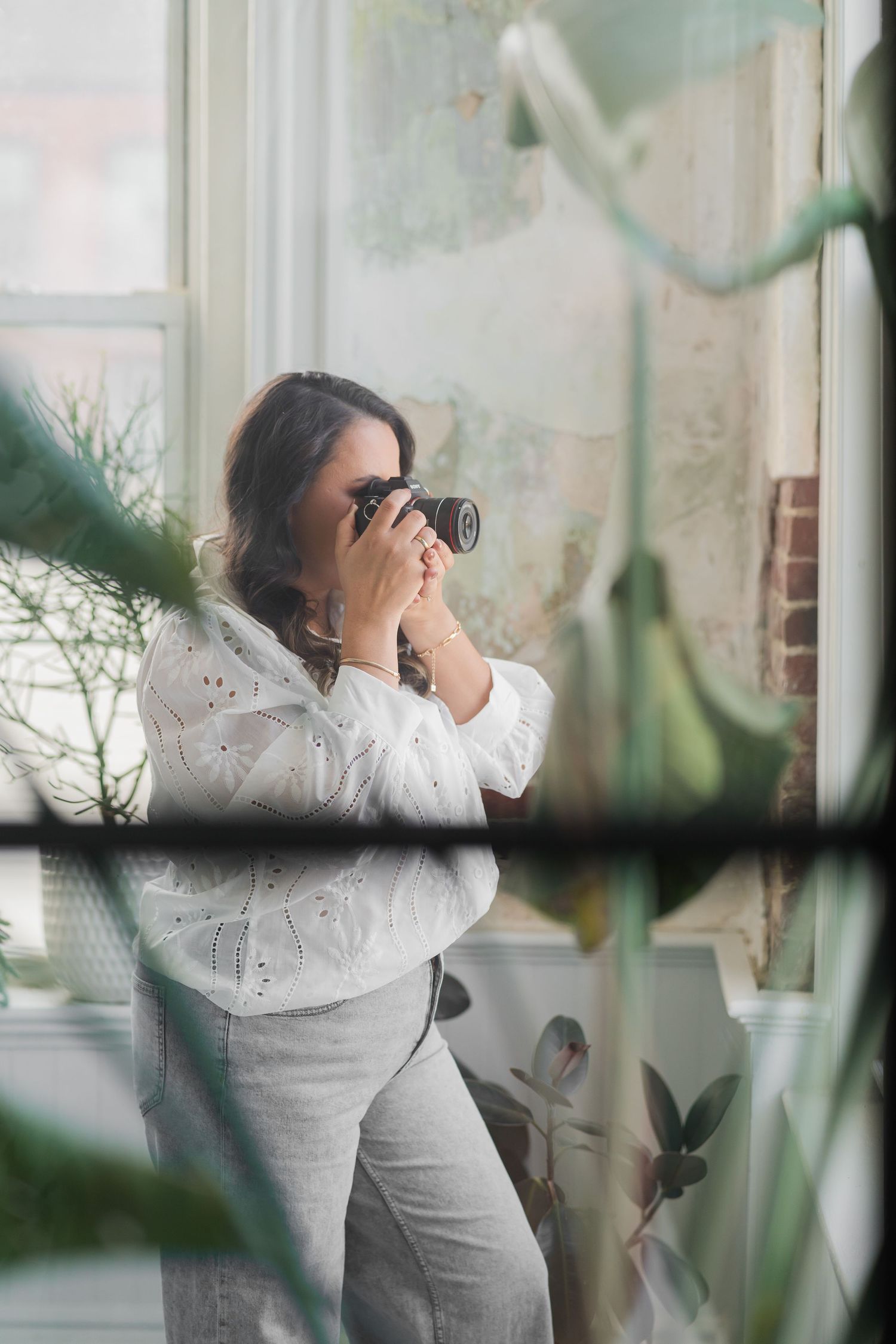 A photographer in a white sweater and gray pants takes pictures through a window surrounded by indoor plants.