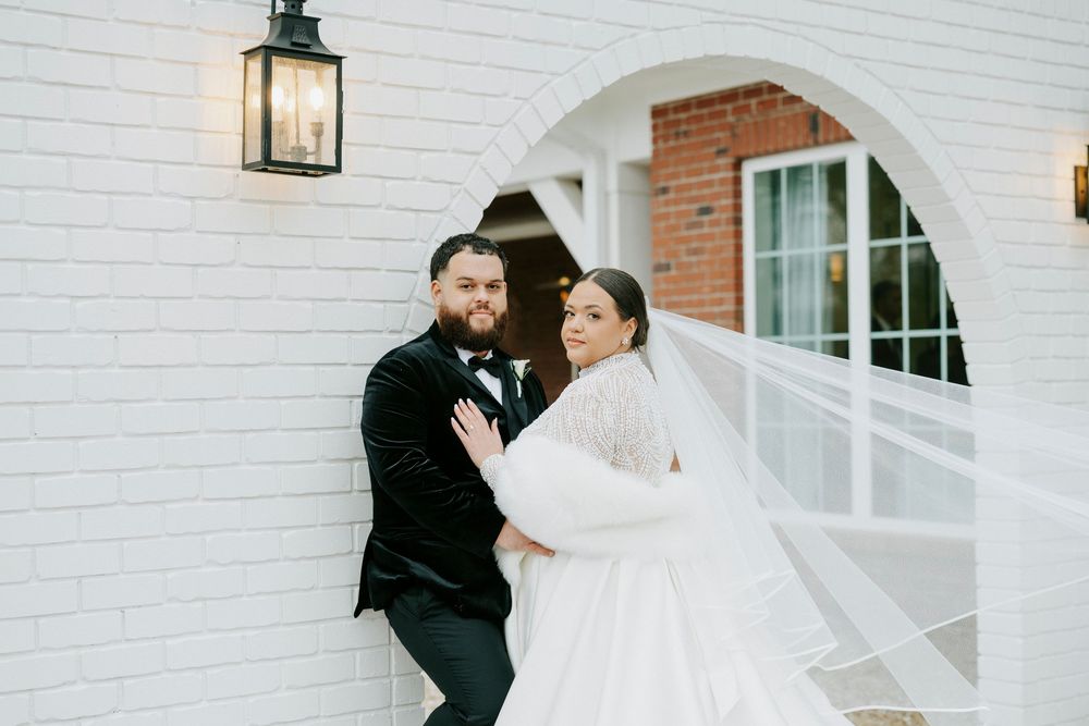 A couple shares a tender moment on their wedding day outside a white brick building with arched windows and lanterns.