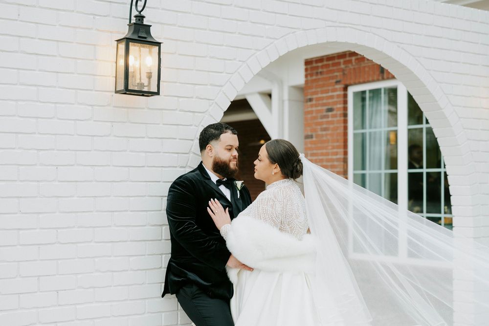 A couple in wedding attire embraces on a white brick patio with arched windows and a lantern in the background.