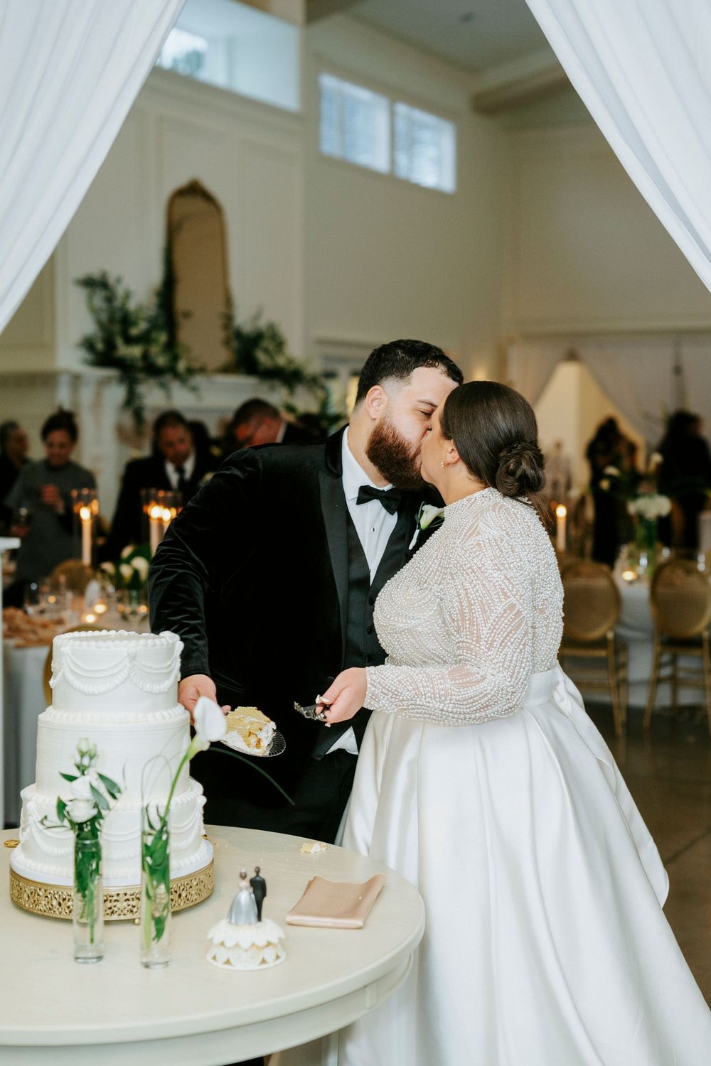 Wedding couple shares intimate moment while cutting their elegant white tiered cake at indoor reception venue.