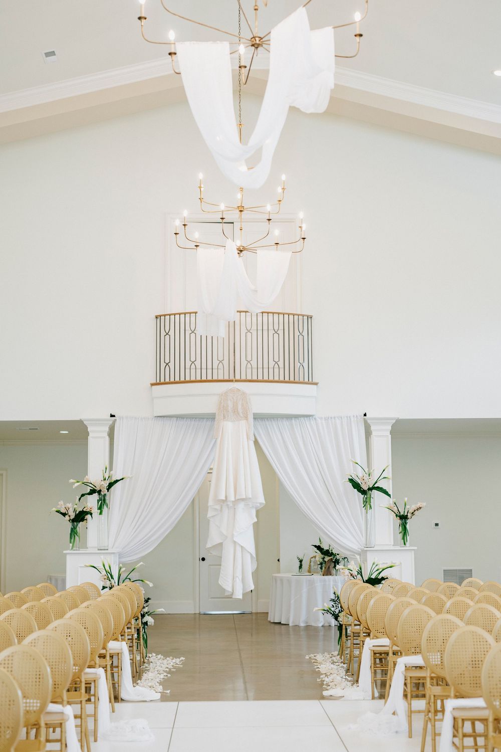 Elegant white wedding ceremony setup with gold chairs, flowing drapes, and chandeliers in a bright indoor venue.