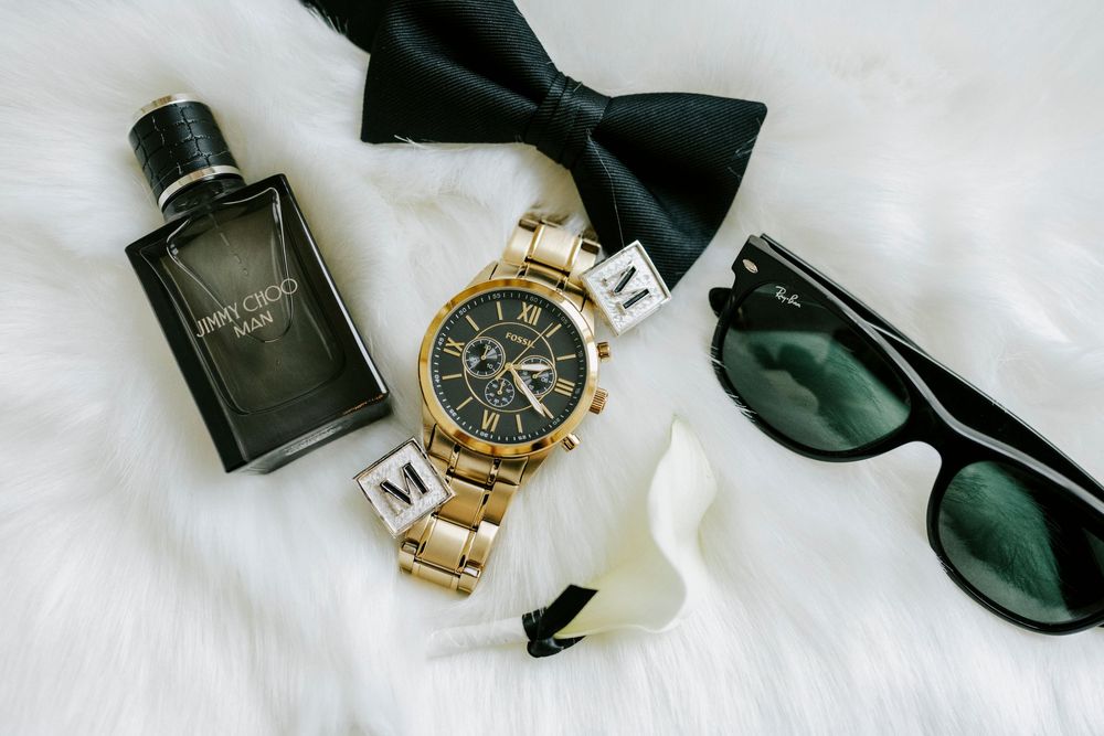 Elegant wedding accessories including gold watch, black bow tie, and cologne bottle arranged on white surface.