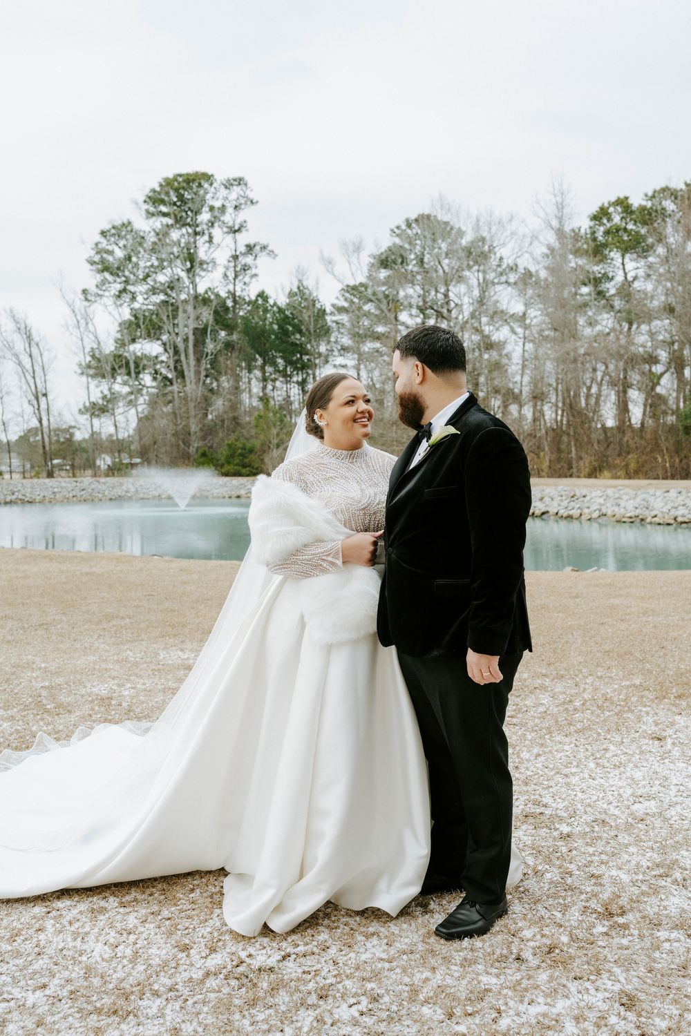 A couple shares a romantic moment in their wedding attire on a snowy field with pine trees in the background.