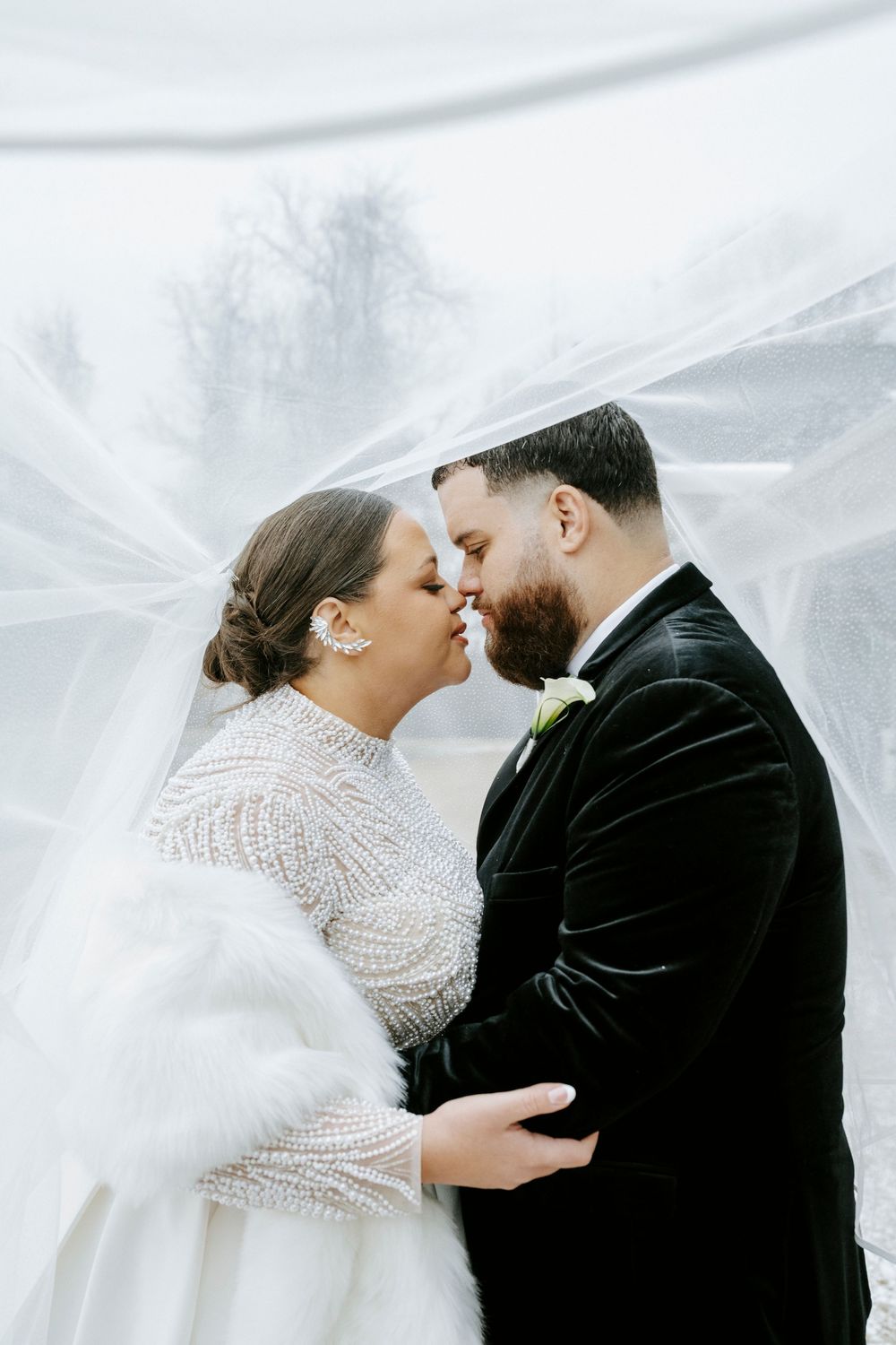 Romantic winter wedding portrait with flowing veil and formal attire against snowy backdrop.