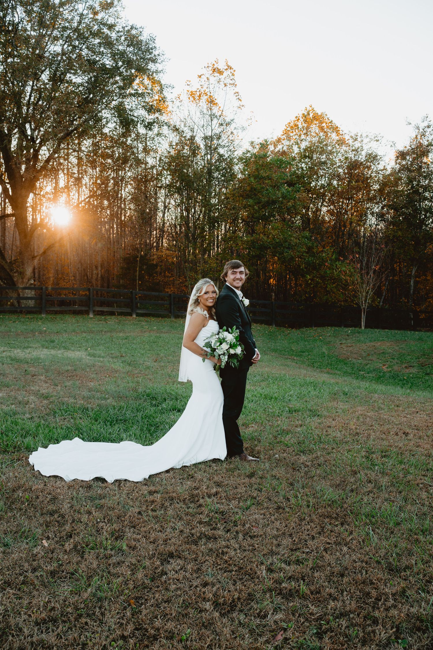 A woman in a white off-shoulder dress poses outdoors against a backdrop of autumn foliage with golden light.