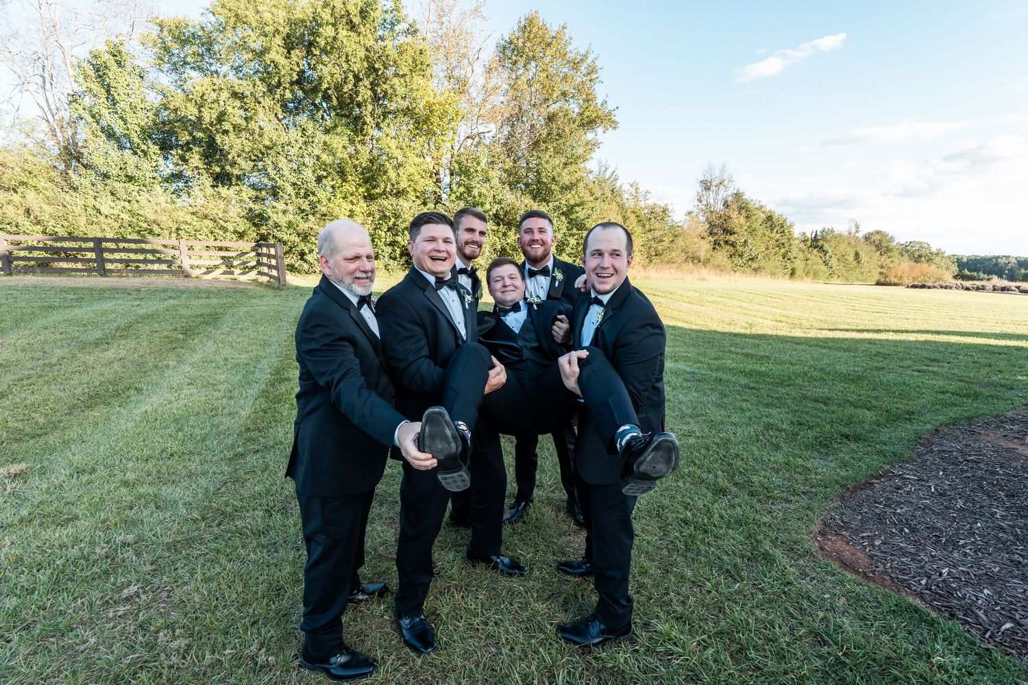 A group in formal black suits and bowties pose together outdoors on a grassy hillside.