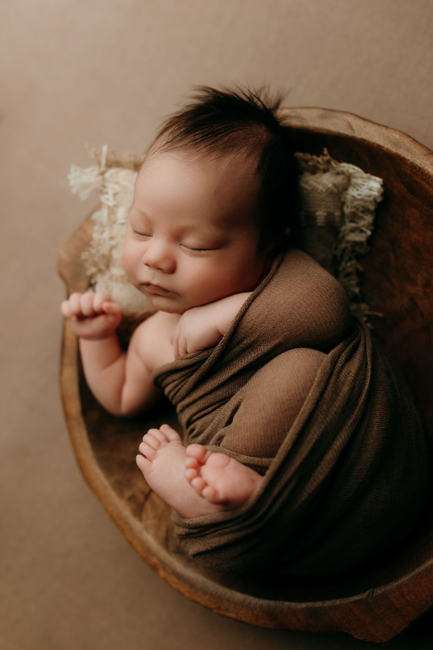 Sleeping newborn wrapped in brown fabric lying in wooden bowl with lace pillow during professional photo session.