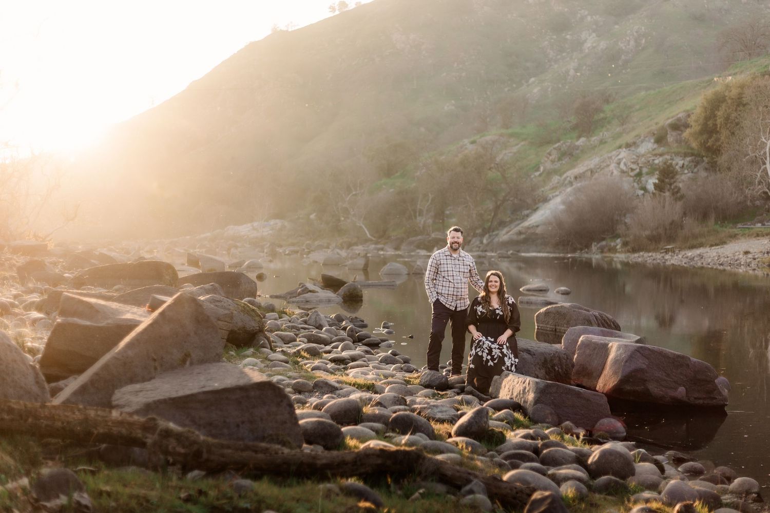 A couple stands together on rocky shoreline during golden hour at a serene mountain lake.