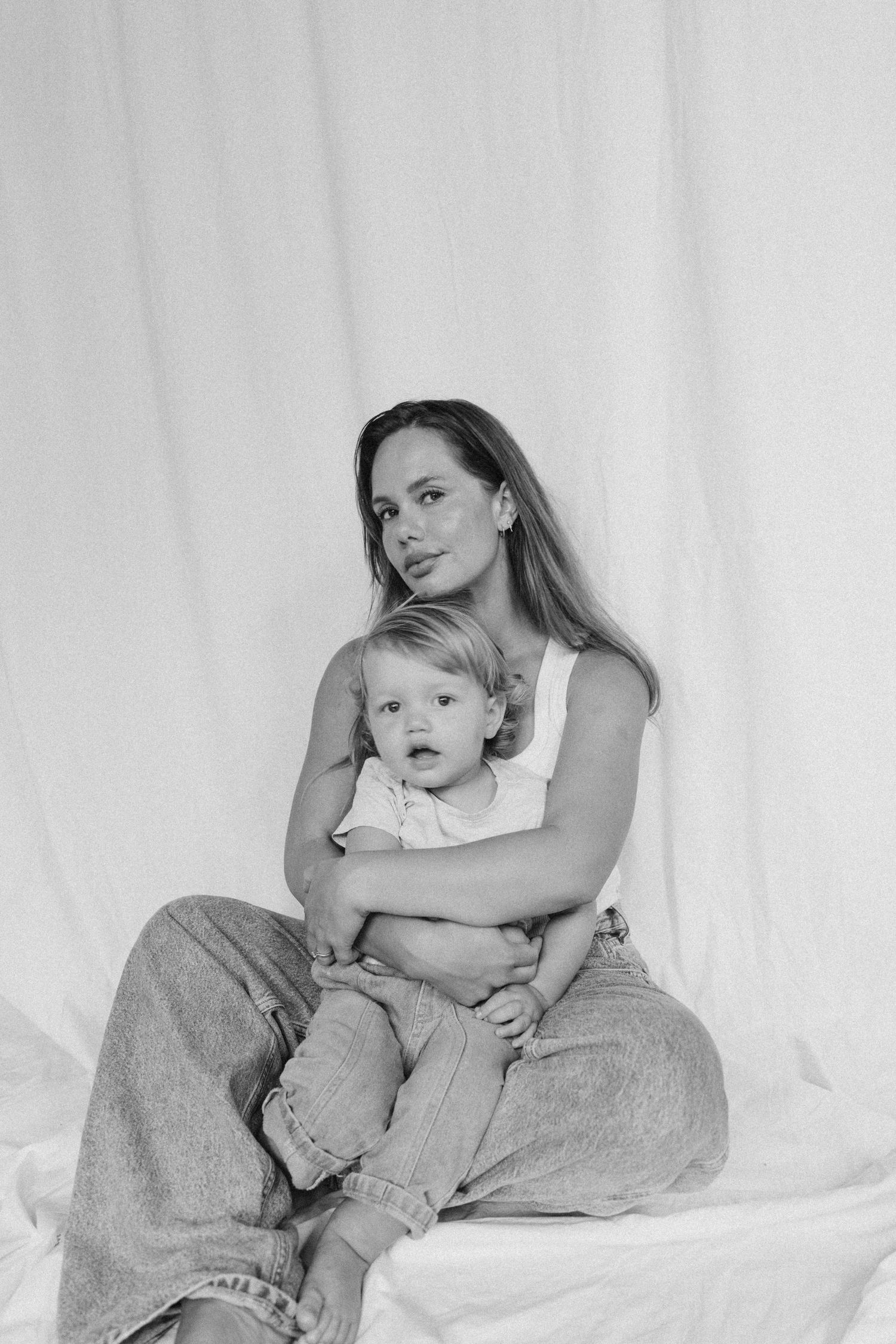 Black and white portrait of mother in white top and jeans holding toddler while seated against white curtain backdrop in New Zealand.