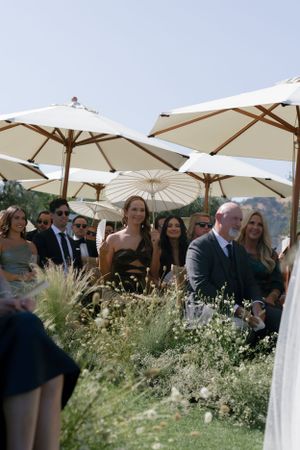 Wedding guests seated outdoors under elegant white parasols surrounded by wildflowers.
