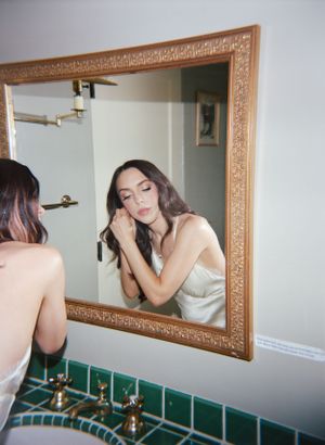 Reflection in an ornate framed mirror shows a person applying makeup at a bathroom counter with green tiles.