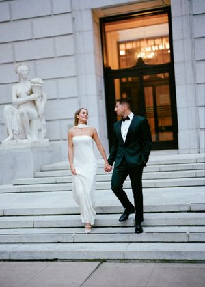 Couple in wedding attire posing on grand stone steps of a classical building.