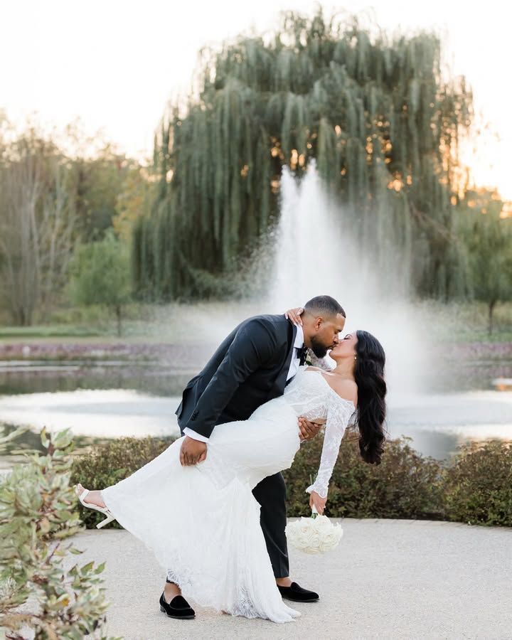 A romantic dip pose in front of a weeping willow tree and fountain during sunset at an outdoor wedding.