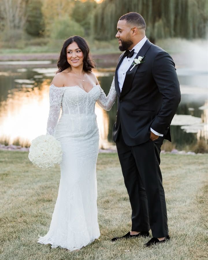 A couple poses together outdoors by a lake at sunset, with the bride in a white lace gown and groom in a black tuxedo.